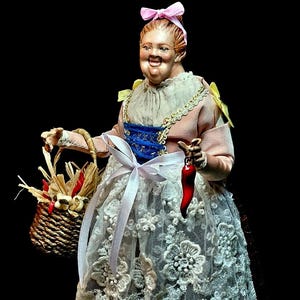 Peut inclure: Figurine détaillée d'une femme en robe rose avec une jupe en dentelle blanche, tenant un panier de maïs et un piment rouge. La femme a un nœud rose dans les cheveux et un corset bleu. La figurine est sur fond noir.