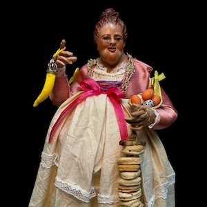Peut inclure: Une figurine représentant une personne aux cheveux bruns en chignon, vêtue d'une veste rose et d'une jupe crème à bordure en dentelle. Elle tient une breloque jaune en forme de corne, un petit panier d'œufs orange et tachetés, et une guirlande de biscuits.