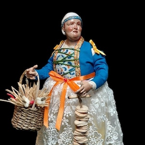 Peut inclure: Figurine d'une femme en costume traditionnel, tenant un panier tressé et un support en bois avec des objets empilés. Elle porte une veste bleue, une jupe en dentelle blanche et un couvre-chef blanc avec une bande bleue. Un nœud orange orne sa taille.