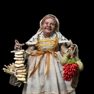 Peut inclure: Une figurine représentant une personne vêtue d'une robe couleur crème avec un châle en dentelle et un couvre-chef. La figurine tient un petit panier avec une pile d'objets plats et clairs d'un côté et un bouquet de piments rouges avec deux courges.