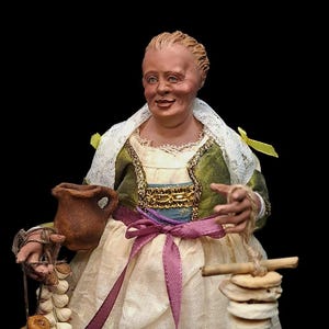 Peut inclure: Figurine détaillée d'une femme en costume traditionnel, tenant une petite cruche marron et une guirlande d'ail. Elle porte un corsage vert et or brodé, un châle en dentelle blanche et une jupe crème. Une pile de petits objets ronds dans sa main.