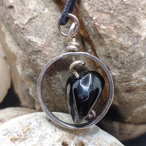 May include: A silver pendant with a black gemstone in a circular frame. The pendant is on a black cord.
