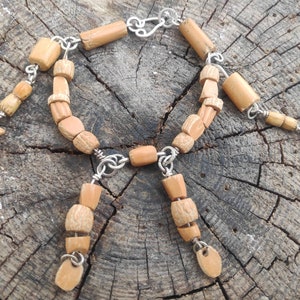 Puede incluir: Un brazalete de cuentas de madera de color beige y marrón con detalles en metal plateado. El brazalete tiene una variedad de formas y tamaños de cuentas, incluyendo cuentas redondas, ovaladas y rectangulares. El brazalete está enhebrado en una cadena de metal plateado.