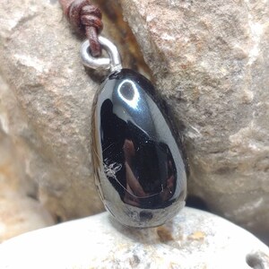 Peut inclure: Un pendentif en pierre précieuse noire sur un cordon en cuir brun. Le pendentif est lisse et brillant, de forme en goutte.
