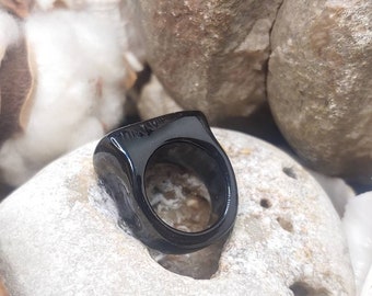 Puede incluir: Un anillo negro brillante de forma rectangular redondeada. El anillo tiene un acabado liso y pulido y una banda ancha. La inscripción "Tiffany & Co." es visible en la parte superior del anillo.