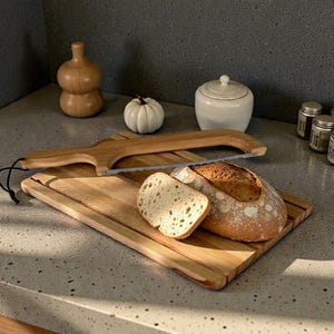 May include: A wooden bread board with a loaf of bread and a slice. A bread saw rests on the board. A small white pumpkin and a decorative wooden jar are in the background.