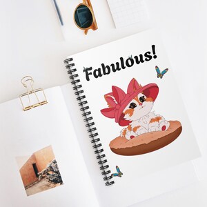 May include: A white spiral notebook with a cartoon cat wearing a pink hat and the word "Fabulous!" in black text. The cat is sitting on a brown pastry with two blue butterflies.