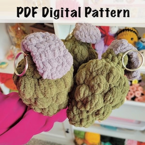 May include: Three crocheted plush keychains shaped like grenades, featuring olive green textured bodies and light purple tops. Each item includes a silver metal keyring attached to the top. The text PDF Digital Pattern is displayed at the top.