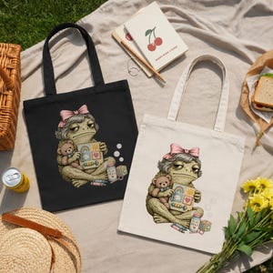 Peut inclure: Un sac fourre-tout noir et un sac fourre-tout blanc, chacun orné d'une illustration graphique d'une grenouille aux cheveux gris avec un nœud rose, tenant un ours en peluche et un bloc jouet, avec un petit flacon étiqueté Play Heals.