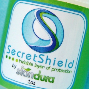 Pode incluir: Close-up de um rótulo de produto com as palavras "SecretShield" e "Invisible layer of protection" em azul e verde. A marca "skindura" também é visível, juntamente com "1 oz". O rótulo tem um fundo branco com uma borda verde.
