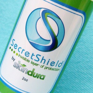 Könnte beinhalten: Eine grüne und weiße Flasche mit dem Text "SecretShield" und "Invisible layer of protection" von "skindura". Das Etikett zeigt ein blaues und grünes abstraktes Design. Die Flasche enthält 59 ml Produkt.
