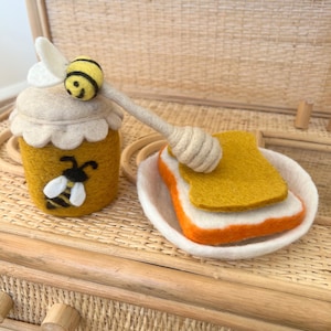 Op de afbeelding: Een vilten honingpot met een bijenontwerp, een honingdipper en een sneetje toast met honing. De honingpot is geel met een wit deksel en een zwart-witte bij. De toast ligt op een wit bord.