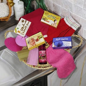 Op de afbeelding: Een rieten mand gevuld met verschillende bad- en schoonheidsproducten, waaronder een roze badborstel, een roze badhandschoen, een blauwe zeep, een fles rozenwater, een kam en verschillende zepen met Arabische tekst.