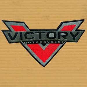 May include: A motorcycle patch featuring the word "VICTORY" in dark gray, with "MOTORCYCLES" below. The design includes a red and gray V-shaped emblem. The patch is on a tan background.