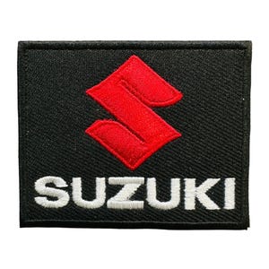 May include: Black embroidered patch featuring the Suzuki logo. The logo is a stylized red 'S' shape, centered on the patch. The word 'SUZUKI' is embroidered in white below the logo. The patch has a black border.