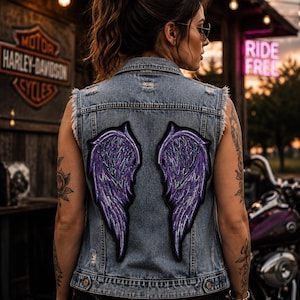 May include: A light blue denim vest featuring large, embroidered purple angel wings on the back. The vest has a distressed finish with frayed armholes, a classic collar, and metal button closures at the waist, showcasing detailed stitching.