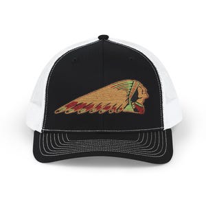 May include: A black and white trucker cap featuring a black front panel with an embroidered graphic of a profile of a person wearing a feathered headdress in shades of tan, red, and green. The cap has a white mesh back and a black curved brim.