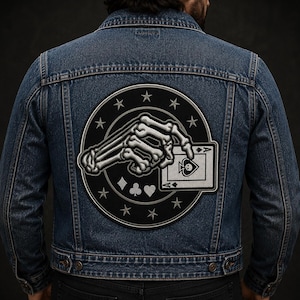 May include: A dark blue denim jacket featuring a large circular embroidered patch on the back. The patch displays a skeletal hand holding an ace of spades playing card, surrounded by a border of stars and the four classic card suit symbols.