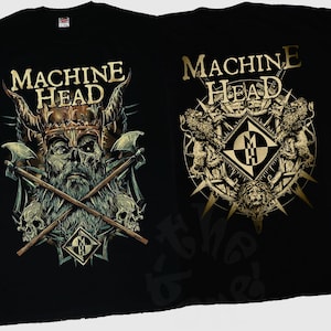 Puede incluir: Dos camisetas negras con gráficos dorados y grises. Una camiseta presenta una calavera con un casco con cuernos, hachas cruzadas y el texto "MACHINE HEAD". La otra camiseta tiene un diseño circular con el nombre y el logotipo de la banda.