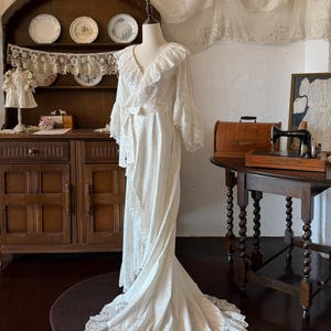 Peut inclure: Une robe portefeuille couleur crème arrivant au sol, dotée d'un décolleté en V avec un large volant, de manches longues évasées et d'une ceinture à nouer assortie. Le tissu présente un motif floral ton sur ton et une bordure en dentelle.
