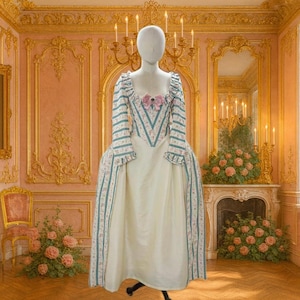 Puede incluir: Vestido de estilo histórico sobre un maniquí. El vestido presenta una falda color crema y un corpiño con mangas a rayas azules y blancas y detalles florales. Un lazo rosa adorna el escote. El fondo es una habitación con detalles dorados y una chimenea.