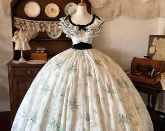 May include: A floor-length white gown featuring a green floral print, a dark green velvet waistband, and a wide neckline with layered ruffled trim. The dress has short sleeves and a full, voluminous skirt that flares out from the waistline.