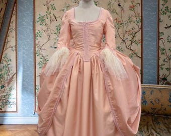 May include: A light pink historical dress on a mannequin. The dress features a square neckline, long sleeves with white lace cuffs, and a full skirt. The bodice is fitted with vertical seams. The dress is displayed in a room with floral wallpaper.