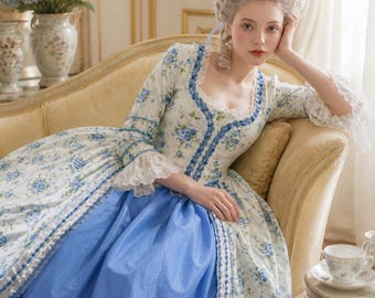 May include: A white and blue floral print dress with a blue skirt, lace trim, and long sleeves. The dress has a square neckline and is reminiscent of historical fashion. The woman is wearing a wig with a blue feather.