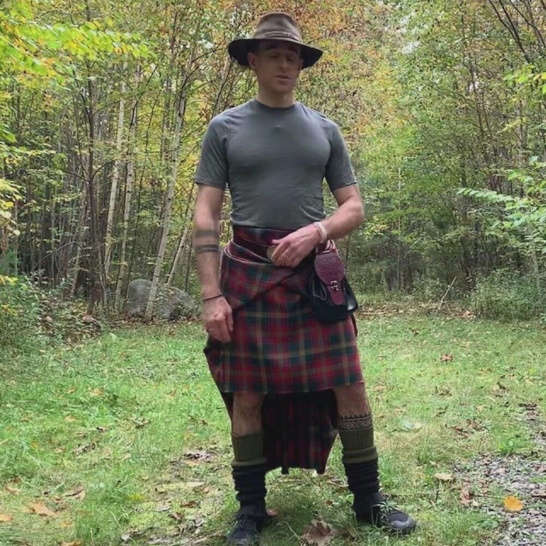 Puede incluir: Una persona en un bosque con un sombrero marr&oacute;n, camiseta gris y un kilt escoc&eacute;s tradicional. El kilt presenta un patr&oacute;n de cuadros rojos, verdes y dorados. Accesorios: un sporran de cuero, calcetines estampados y zapatos negros.