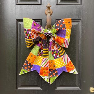May include: A large decorative bow made from a patchwork of fabric squares featuring Halloween themes. The patterns include orange pumpkins, black bats, candy corn, purple spiderwebs, and green polka dots in a vibrant, multi-coloured design.