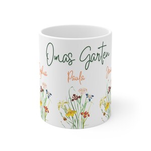 Könnte beinhalten: Weiße Keramiktasse mit einem grün-schwarzen Design, auf dem "Omas Garten" und "Paula" in Orange stehen. Die Tasse ist mit einem Aquarell-Blumenmuster aus bunten Blumen und grünen Blättern verziert.