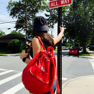 May include: A red drawstring backpack with a front pocket is worn by a person standing near a stop sign. The stop sign is red with white lettering and the words "ALL WAY" below. A black cap is worn.