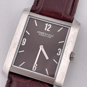 Könnte beinhalten: Eine rechteckige Kenneth Cole Reaction Armbanduhr mit braunem Zifferblatt und braunem Lederarmband. Die Uhr hat ein silberfarbenes Gehäuse und zeigt die Zahlen 12, 3, 6 und 9 an. Die Zeiger der Uhr sind silberfarben.