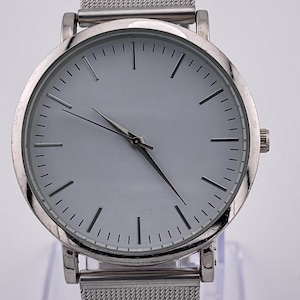 Puede incluir: Reloj de pulsera plateado con una correa de malla y un diseño minimalista. El reloj presenta una esfera redonda de color claro con manecillas negras y marcadores de hora sencillos. La correa es de metal fino tejido.