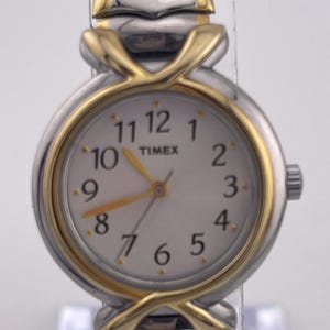 Puede incluir: Un reloj Timex con una correa y caja de metal plateado y dorado. La esfera del reloj es blanca con números negros y agujas doradas. El reloj tiene el logotipo de Timex.
