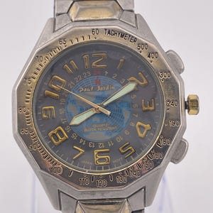 May include: Silver and gold-toned Paul Jardin quartz watch with a dark gray face. The watch features a world map design, luminous hands, and a tachymeter bezel. The watch band is silver and gold-toned.