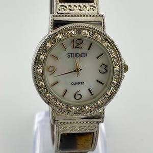 Puede incluir: Reloj de cuarzo Studio Time plateado con esfera blanca, manecillas doradas y bisel con cristales. El reloj tiene una correa decorativa con detalles marrones y dorados. La esfera muestra los números 12, 3, 6 y 9.