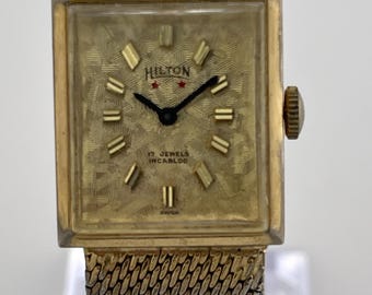 Reloj de vestir rectangular vintage Hilton, década de 1960, con pulsera de malla dorada.
