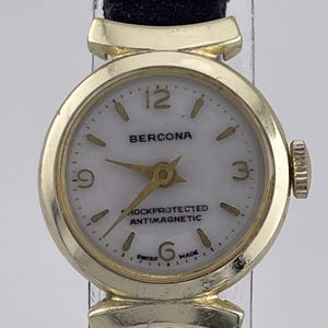 Könnte beinhalten: Eine vintage goldfarbene BERCONA Armbanduhr mit weißem Zifferblatt und schwarzem Armband. Das Zifferblatt der Uhr hat goldene Zeiger und Markierungen mit dem Text "SHOCKPROTECTED ANTIMAGNETIC" und "SWISS MADE". Eine klassische Uhr.