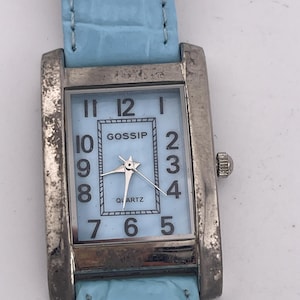 May include: A rectangular silver-toned watch with a light blue face and matching band. The watch face displays the word "GOSSIP" and the word "QUARTZ". The watch has a light blue leather-like band.