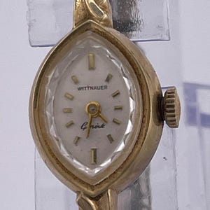 May include: A vintage gold-toned Wittnauer watch with a unique oval face. The watch features a white dial with gold-colored hour markers and hands. The watch band is a delicate gold chain. The watch face reads "Wittnauer" and "Gnote".