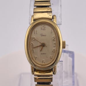 Pode incluir: Um relógio Timex a quartzo dourado com mostrador oval e pulseira extensível. O mostrador do relógio é de cor bege claro com algarismos e ponteiros pretos. O relógio é exibido em um suporte de plástico transparente.