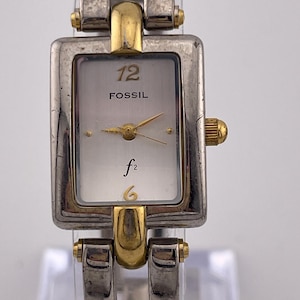 May include: A Fossil brand wristwatch with a rectangular silver-tone face and gold-tone accents. The watch face displays the number 12, the brand name, and the time hands. The watch band is silver-tone with gold-tone details.
