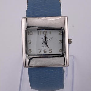 Könnte beinhalten: Eine silberfarbene Armbanduhr mit rechteckigem Zifferblatt und hellblauem Armband. Das Zifferblatt ist weiß mit schwarzen Ziffern und Zeigern. Das Armband ist strukturiert und scheint aus Leder zu sein.