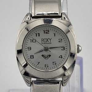 May include: A silver Roxy Quiksilver watch with a light-coloured face. The watch has a silver band and a round face with black numerals and hands. The Roxy logo is visible on the watch face.