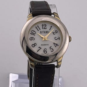 May include: A silver-toned Gitano quartz watch with a white face, black numerals, and gold hands. The watch has a black leather band and gold accents. The watch face displays the word "Gitano".