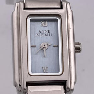 May include: A silver-toned Anne Klein II wristwatch with a rectangular face. The watch face is light blue with silver hands and Roman numeral hour markers. The watch band is also silver-toned and has a clasp closure.