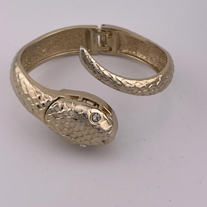 Può includere: Bracciale a forma di serpente color oro con design a cerniera. Il bracciale presenta un motivo strutturato a forma di scaglia e una testa di serpente con una piccola gemma trasparente. Il bracciale è aperto, mostrando il meccanismo di chiusura.