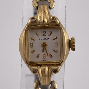 May include: A vintage Bulova wristwatch with a gold-tone case and band. The watch face is square with a white background, gold hands, and black numerals. The watch reads "Bulova" and "Swiss".