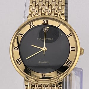 May include: A gold-tone Halston quartz wristwatch with a black face and Roman numeral hour markers. The watch has a gold-tone metal band.